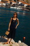 Woman in a navy tank dress with a shell pendant and a beaded necklace holding a raffia bag. 