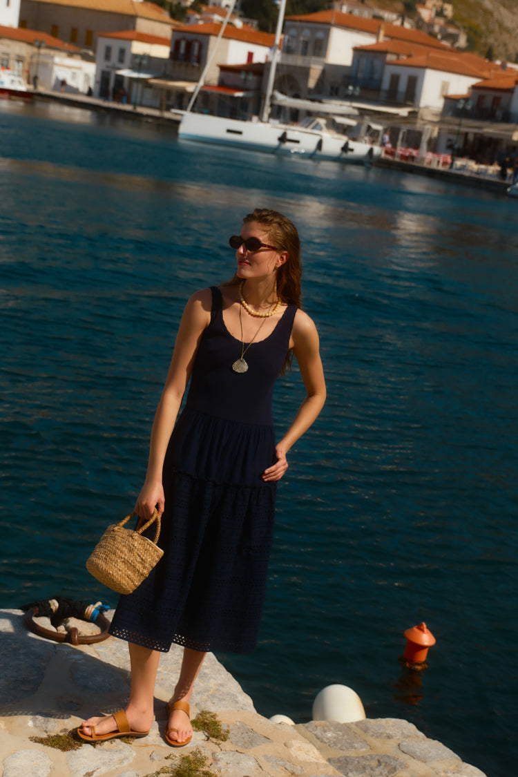 Woman in a navy tank dress with a shell pendant and a beaded necklace holding a raffia bag. 