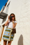 Woman standing in front of white building holding a camera. She wears sunglasses, a green and blue patterned mini skirt, and a white tee. 