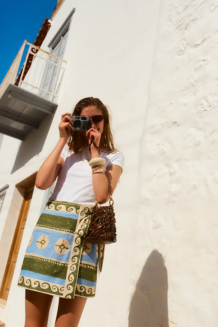 Woman standing in front of white building holding a camera. She wears sunglasses, a green and blue patterned mini skirt, and a white tee. 