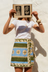 Woman holding an open book in front of her face. She wears a white tee and a green and blue patterned wrap skirt. 