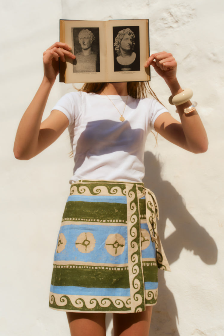 Woman holding an open book in front of her face. She wears a white tee and a green and blue patterned wrap skirt. 