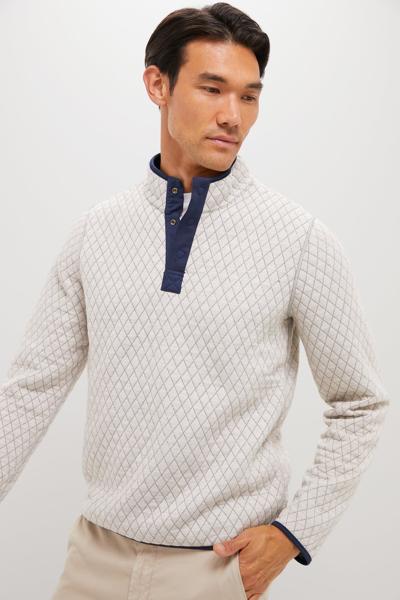 A man stands against a plain background wearing a Marine Layer light-colored quilted pullover with a navy buttoned collar and light beige pants.