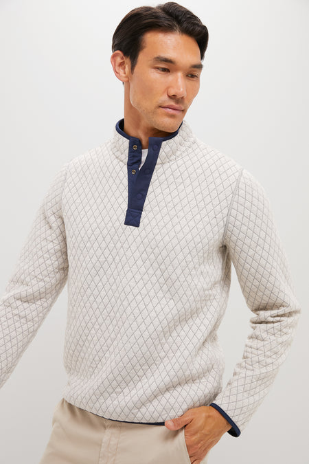 A man stands against a plain background wearing a Marine Layer light-colored quilted pullover with a navy buttoned collar and light beige pants.