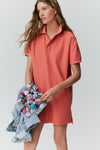 Woman standing in front of a white background in a red polo dress and holding a jean jacket covered in plaid buttons. 