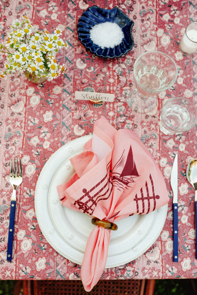Close up of a summer table setting with a red tablecloth, a pink sailboat napkin, and blue silverware. 