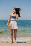 Woman standing in front of the water in a blue stripe one piece swimsuit and white shorts. 