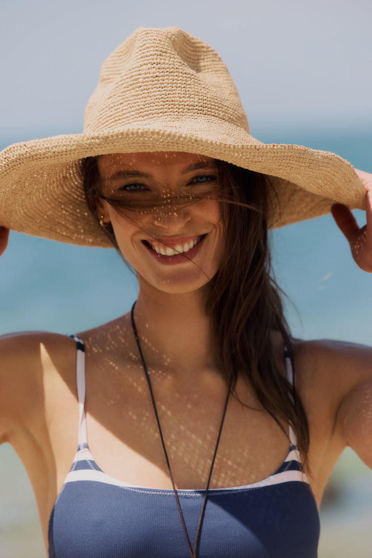 Woman standing outside in the sun in a straw had and blue one-piece swimsuit. 