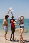 Three people on the beach. Man is in blue swim shorts holding the woman in the middle's towel up on her head. Woman in the middle is in a red swimsuit. Woman to the right is in a blue and white striped swimsuit with a towel on her head. 