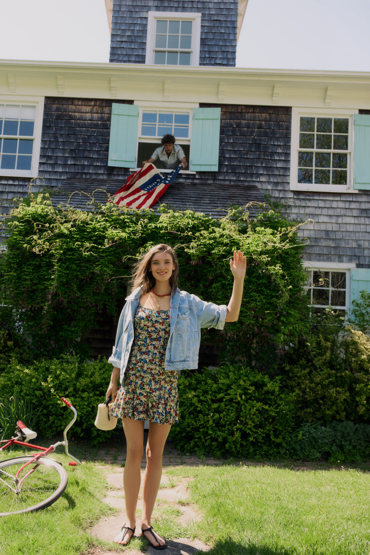 Woman standing in front of a house in a floral dress and jean jacket. 
