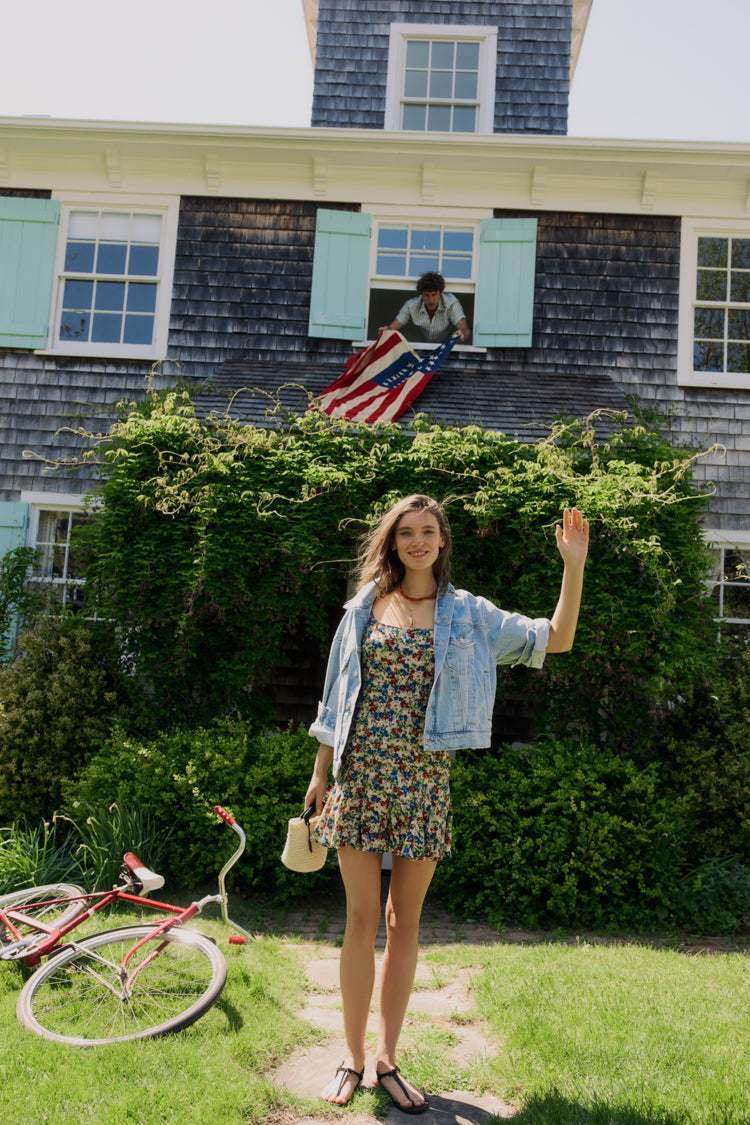Woman standing in front of a house in a floral dress and jean jacket. 