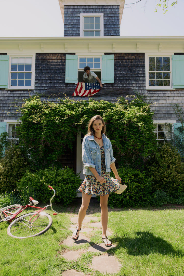 Woman standing in front of a house in a floral dress and jean jacket. 