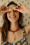 Closeup of woman standing in front of a floral backdrop holding daisies in front of her eyes in a red beaded necklace and fish necklace and a floral dress. 