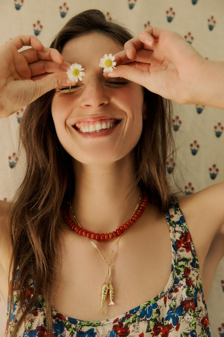 Closeup of woman standing in front of a floral backdrop holding daisies in front of her eyes in a red beaded necklace and fish necklace and a floral dress. 
