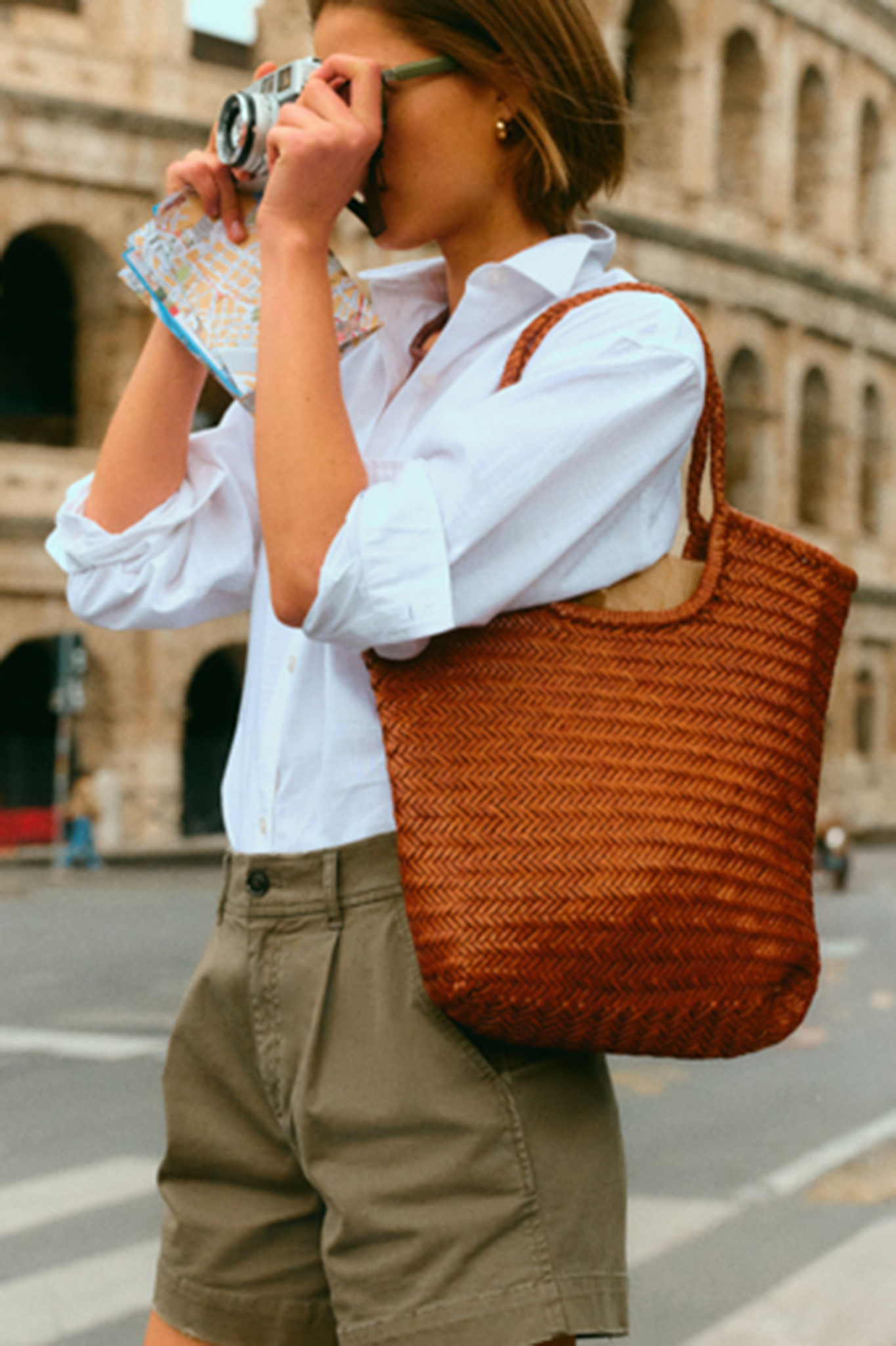 Woman taking a photo in front of the Colosseum in a white button down, olive shorts, and a woven leather bag. 