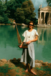 Woman holding a toy sailboat by the water in a white top and a blue skirt. 