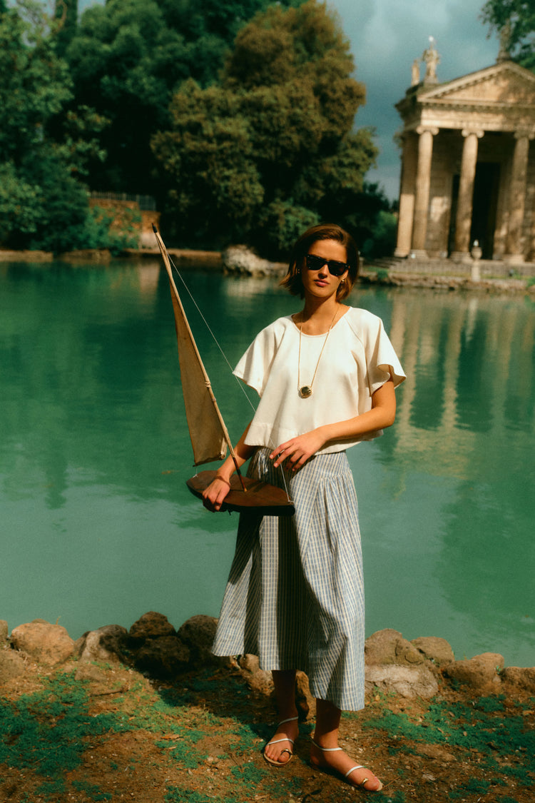 Woman holding a toy sailboat by the water in a white top and a blue skirt. 