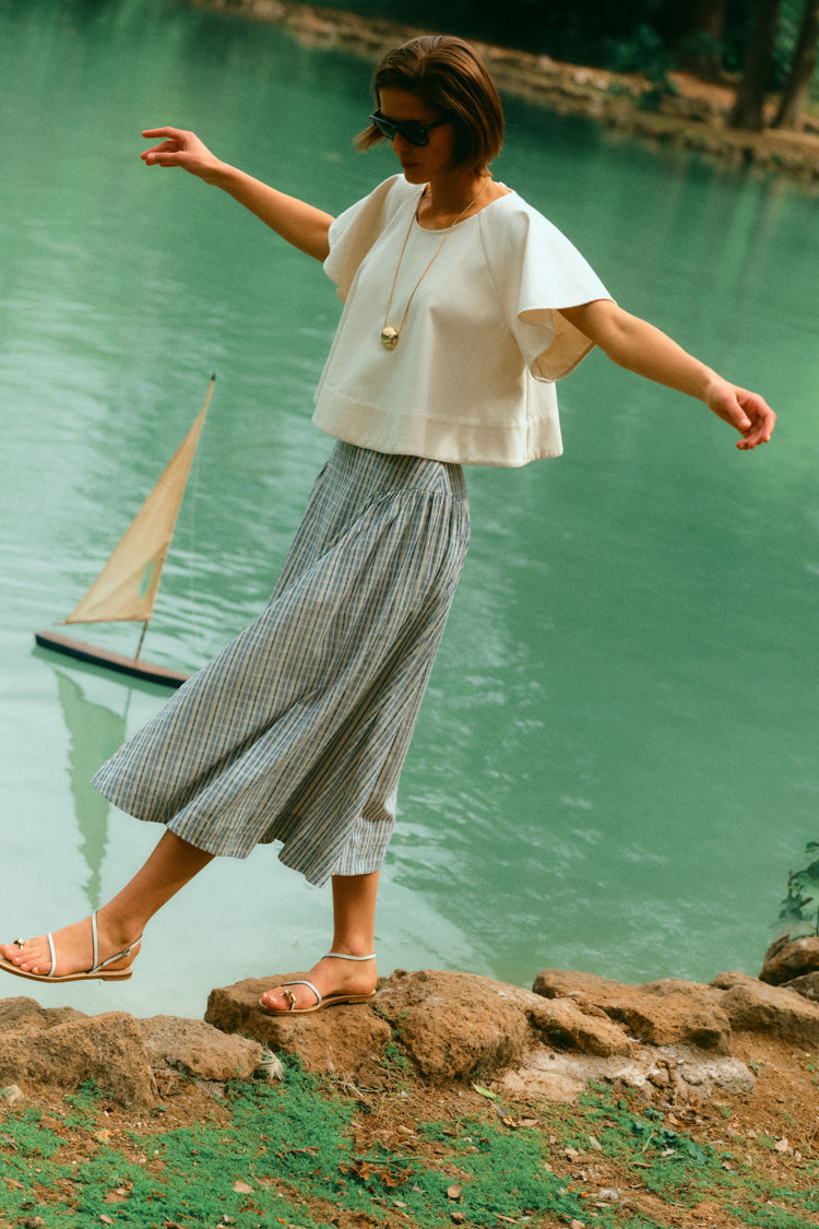 Woman walking on the edge of water in a white top and a stripe skirt. 