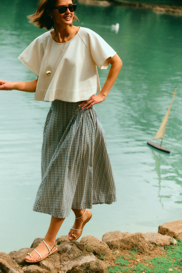 Woman standing by the water in a white top and a blue skirt. 
