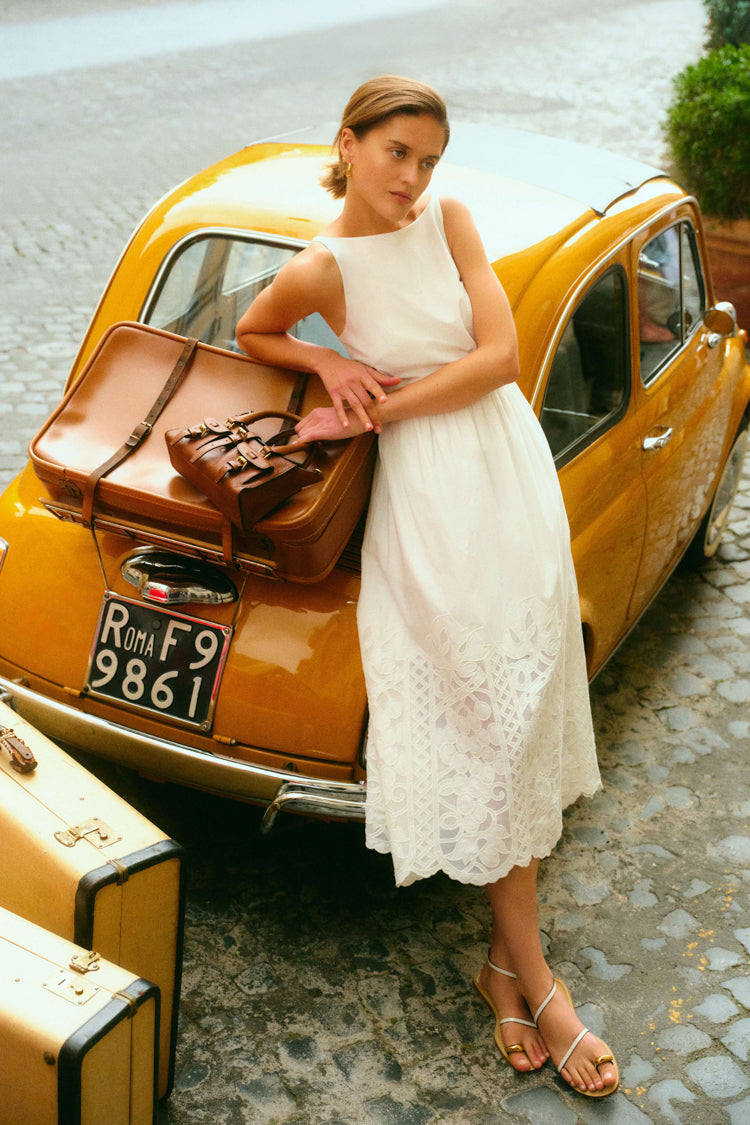 Woman leaning on a yellow taxi and a suitcase in a white dress. 