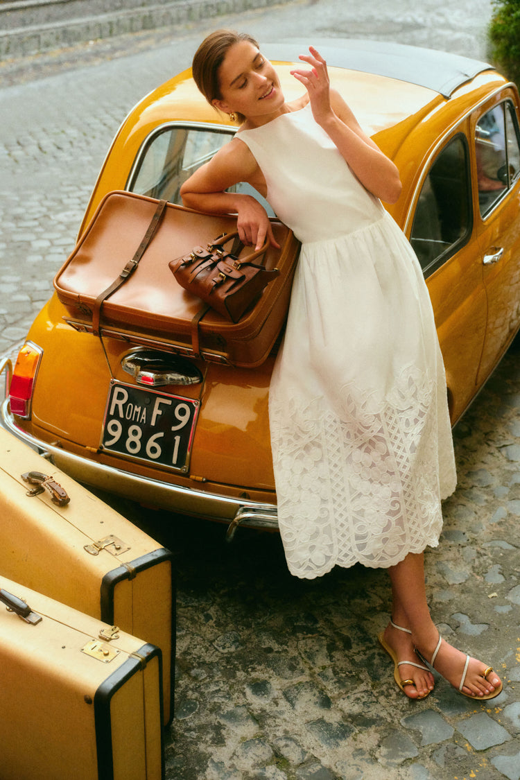 Woman leaning on a yellow taxi and a suitcase in a white dress. 