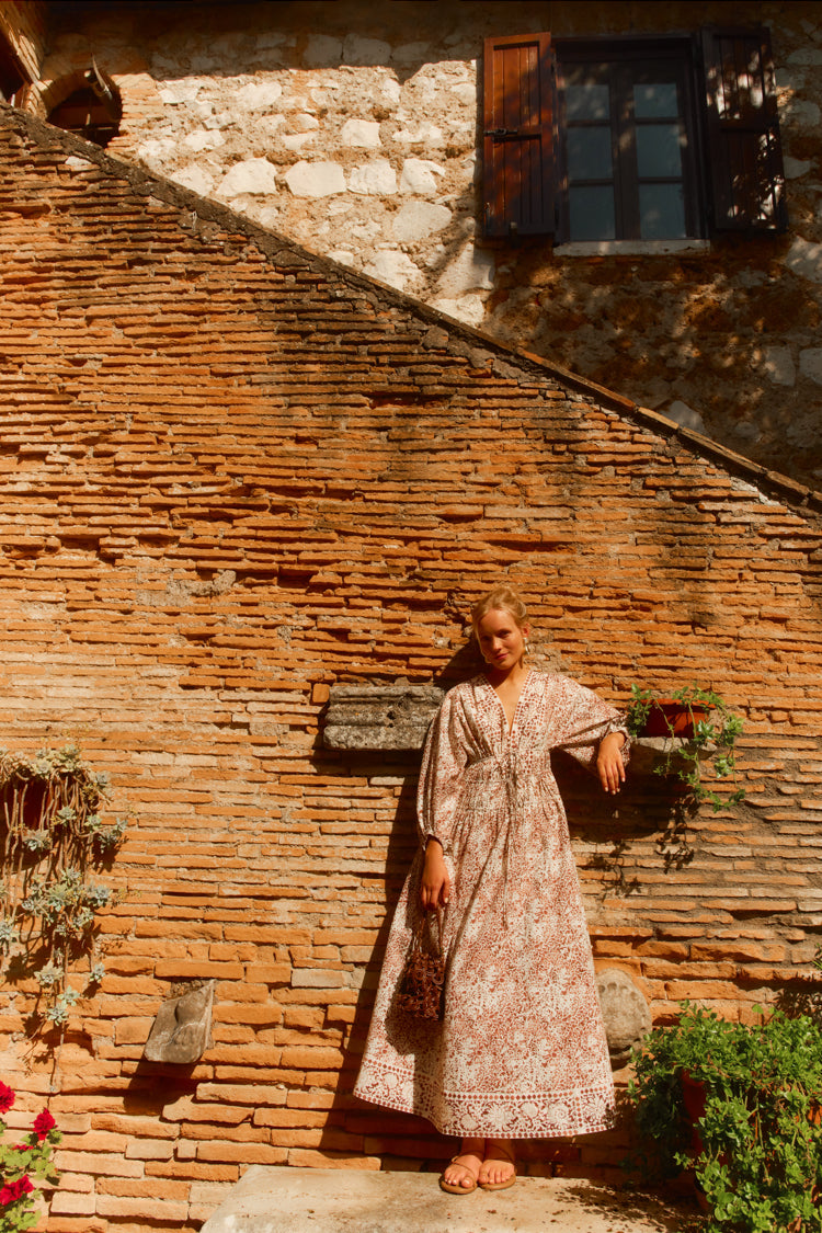Woman standing against a brick wall in a brown and white printed maxi dress. 