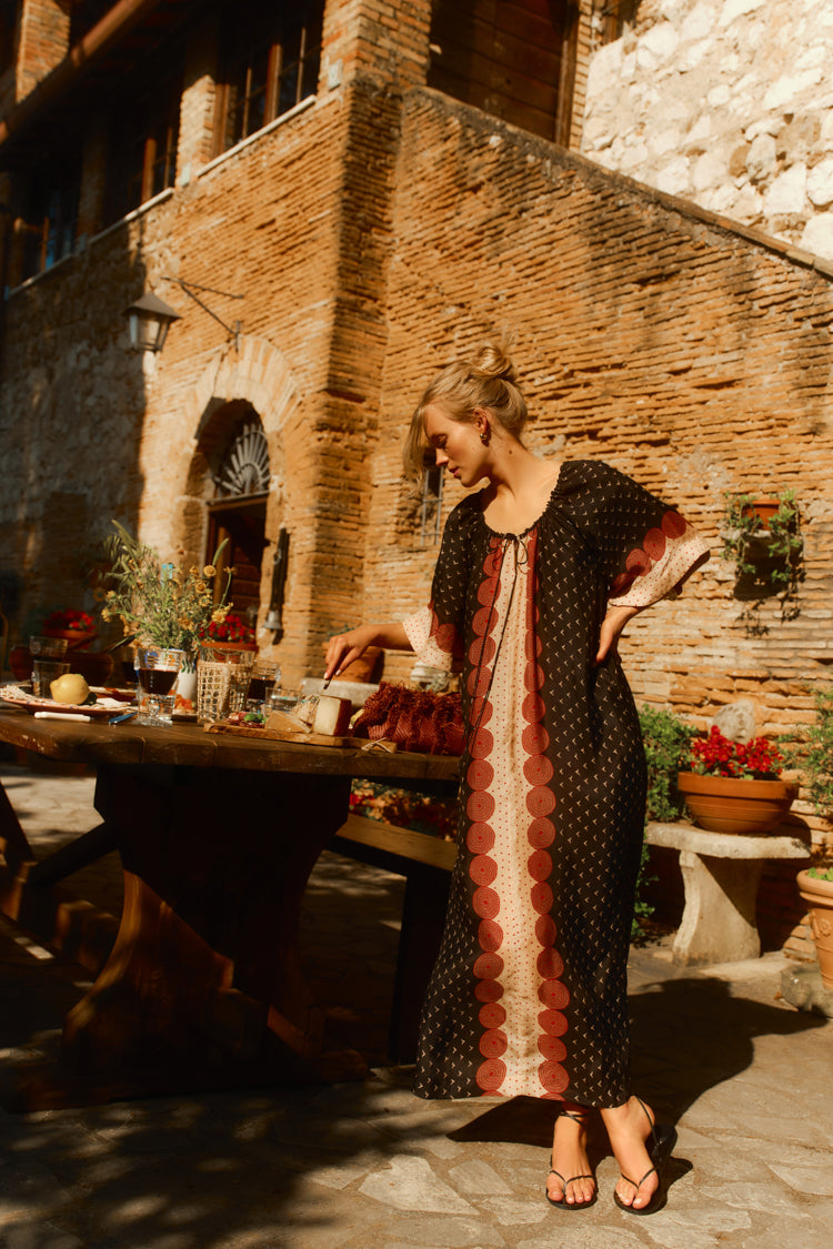 Woman leaning on a table outside of a Tuscan villa in a black and red printed caftan. 