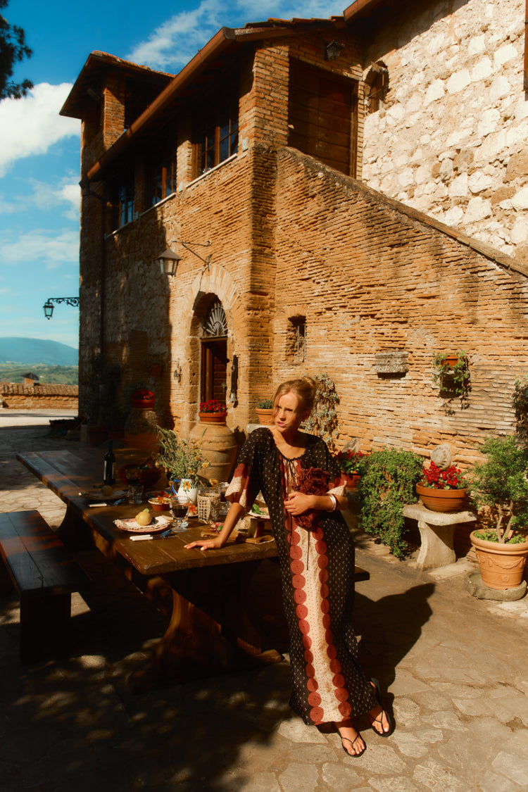 Woman leaning on a table outside of a Tuscan villa in a black and red printed caftan. 