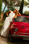 Woman leaning on a red car in a white top and a white lace skirt. 