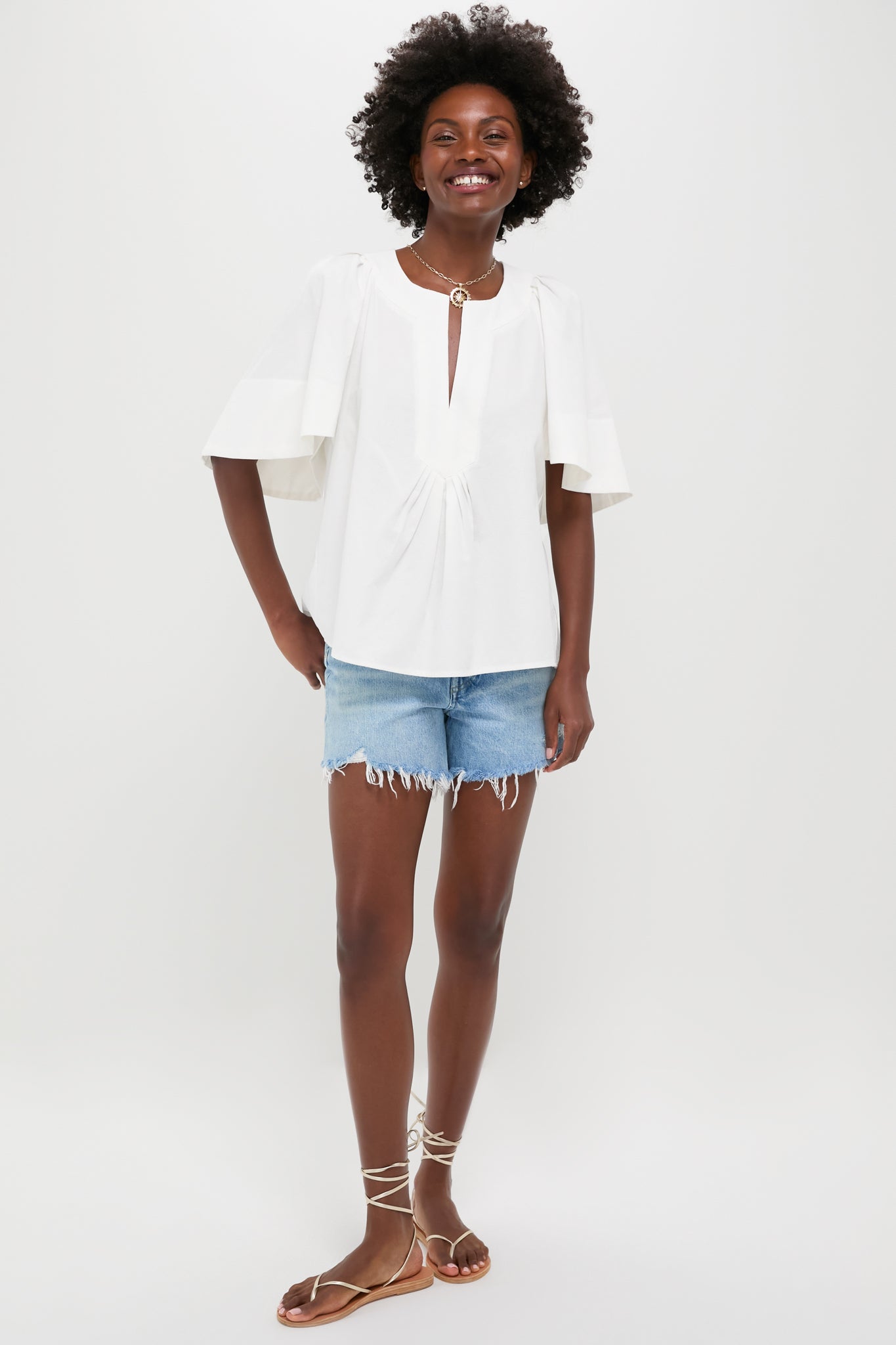 A person stands against a plain background wearing a Pomander Place 100% cotton white V-neck flutter sleeve top, light blue frayed denim shorts, and brown strappy sandals.