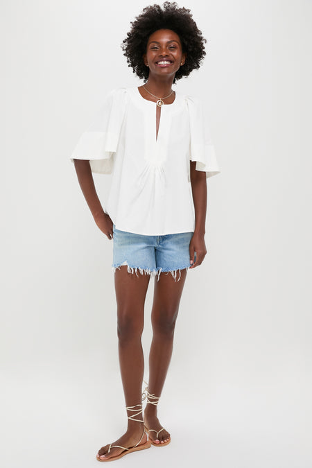 A person stands against a plain background wearing a Pomander Place 100% cotton white V-neck flutter sleeve top, light blue frayed denim shorts, and brown strappy sandals.