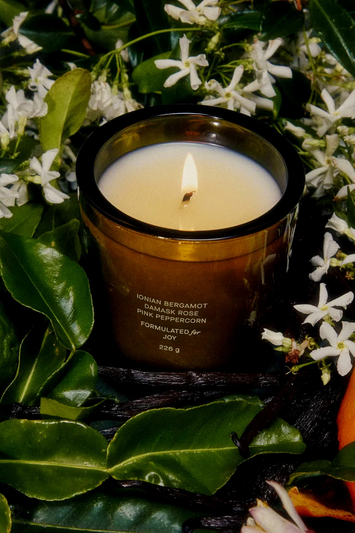 Flamingo Estate’s plant-based candle in a brown glass jar, labeled Ionian Bergamot, Damask Rose, and Pink Peppercorn, is surrounded by green leaves and white flowers and evokes their signature Jasmine fragrance.