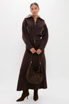 A woman stands against a plain white background wearing CLEA’s dark brown alpaca-blend bomber jacket and matching long skirt, paired with brown boots and a brown suede handbag.