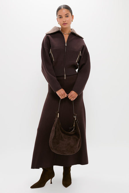 A woman stands against a plain white background wearing CLEA’s dark brown alpaca-blend bomber jacket and matching long skirt, paired with brown boots and a brown suede handbag.