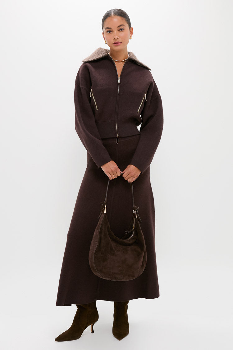 A woman stands against a plain white background wearing CLEA’s dark brown alpaca-blend bomber jacket and matching long skirt, paired with brown boots and a brown suede handbag.