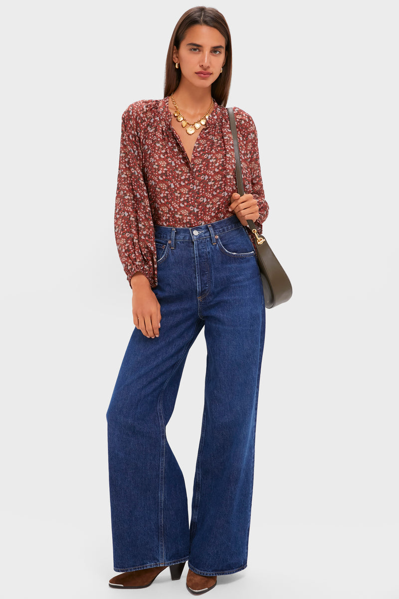 A woman stands against a plain background wearing the Tuckernuck Sawyer Blouse—a red floral print top with a relaxed fit—paired with wide-leg blue jeans, brown boots, a statement necklace, and a shoulder bag.