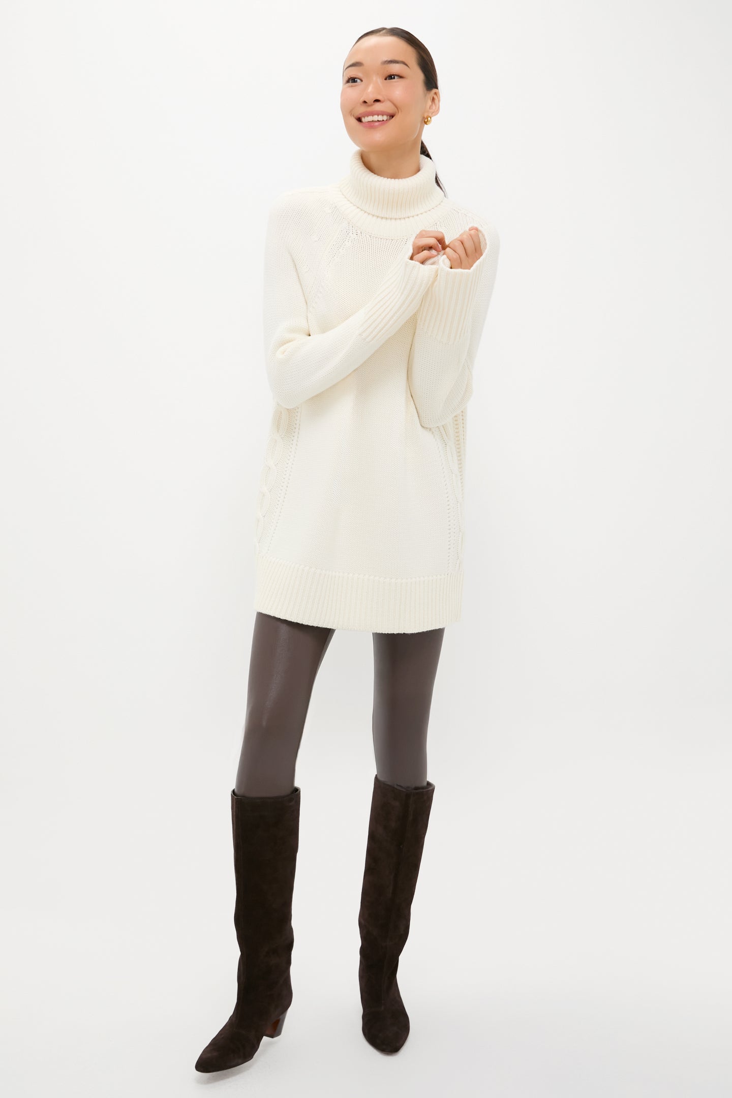 A person models a Tuckernuck long white cable-knit turtleneck sweater with dark leggings and black knee-high boots, standing against a plain white background and smiling with arms slightly raised.