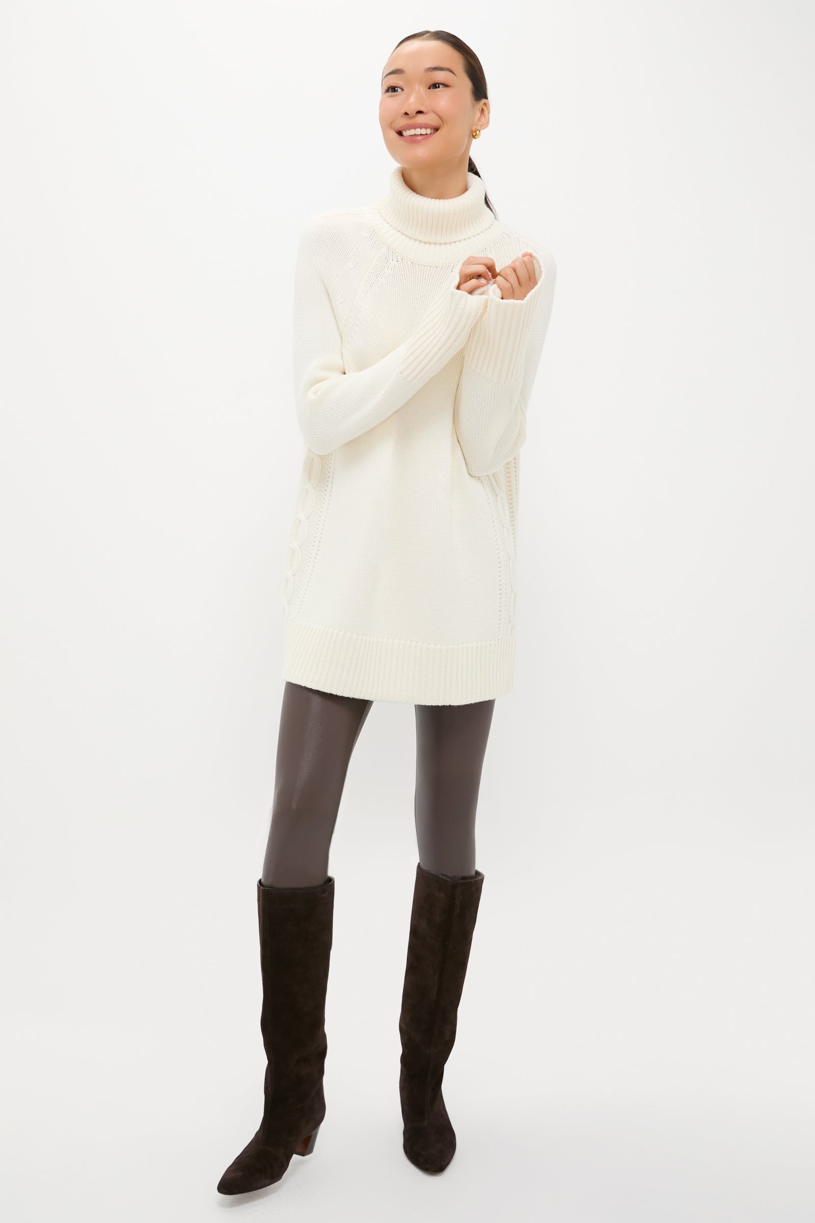A person models a Tuckernuck long white cable-knit turtleneck sweater with dark leggings and black knee-high boots, standing against a plain white background and smiling with arms slightly raised.
