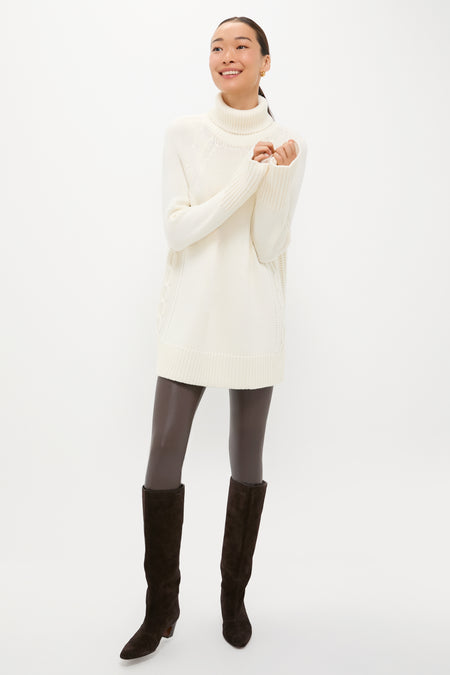 A person models a Tuckernuck long white cable-knit turtleneck sweater with dark leggings and black knee-high boots, standing against a plain white background and smiling with arms slightly raised.