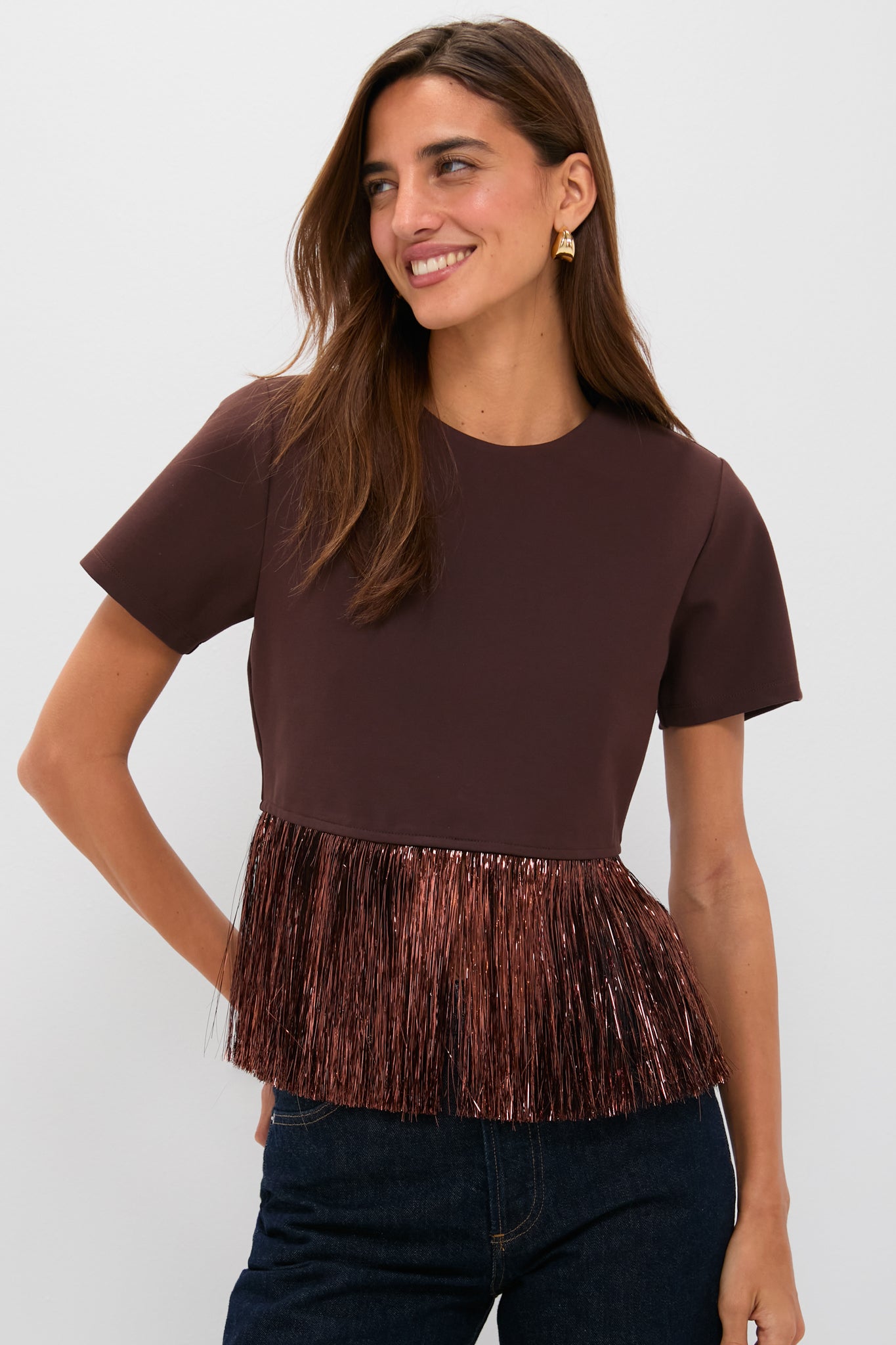 A woman wears a Hyacinth House Francie blouse, a short-sleeve brown party top featuring metallic fringe at the hem, styled with dark jeans.