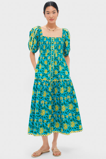A woman faces forward in a Pink City Prints blue and yellow organic cotton floral midi dress with puffed sleeves, a square neckline, and sandals—highlighting sustainable style.