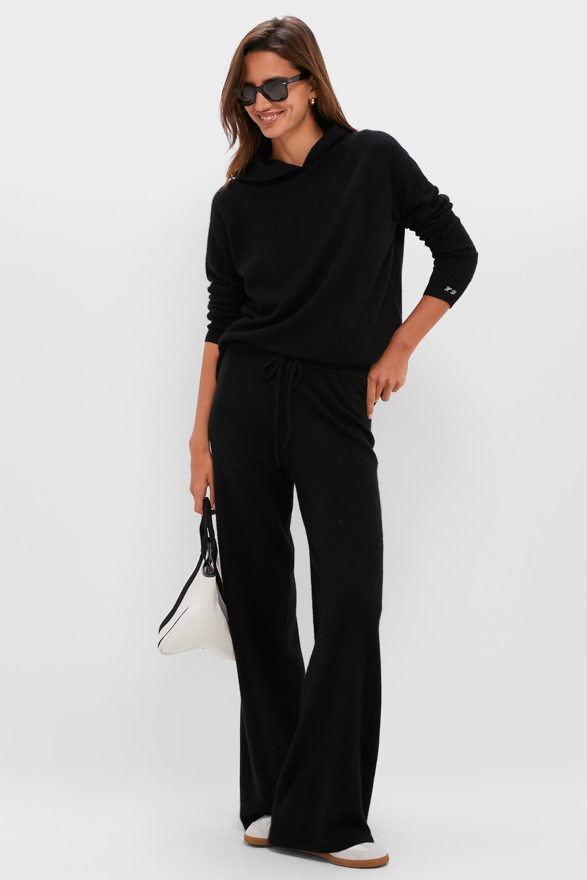 A woman wears a matching black cashmere set with long sleeves and wide-leg pants, styled with sunglasses, a white bag, and light-colored slip-on shoes.