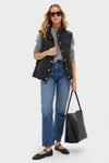 A woman models Tuckernuck's Black Inez Puffer Vest over a gray sweater with blue jeans, black flats, and a large black tote—spotlighting chic women's outerwear against a plain background.