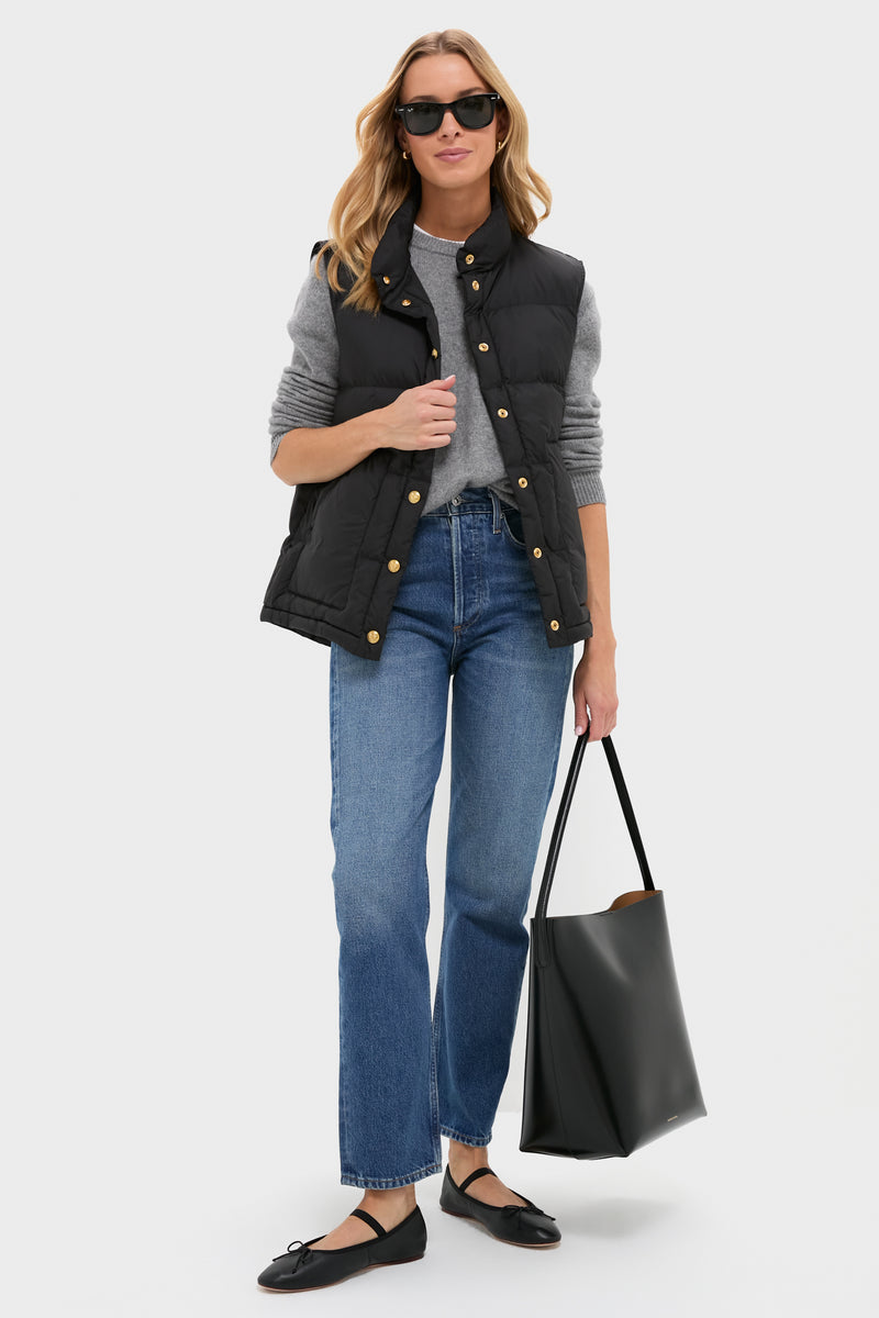 A woman models Tuckernuck's Black Inez Puffer Vest over a gray sweater with blue jeans, black flats, and a large black tote—spotlighting chic women's outerwear against a plain background.