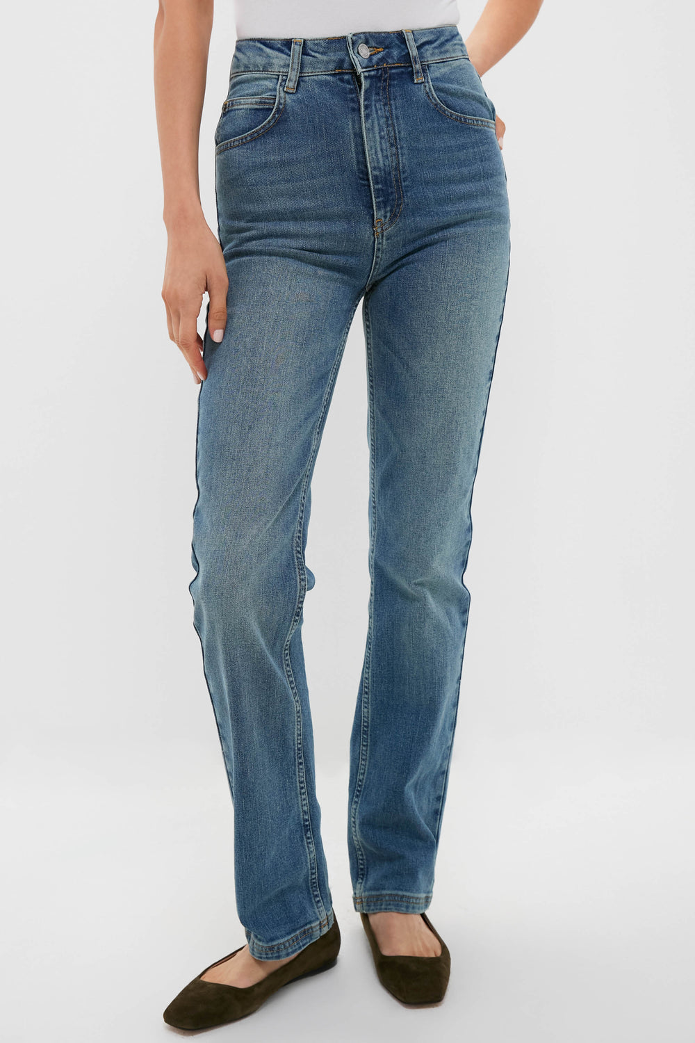 A person wearing high-rise, straight cut Vanessa Bruno jeans and brown flats stands with one hand by their side against a plain background.