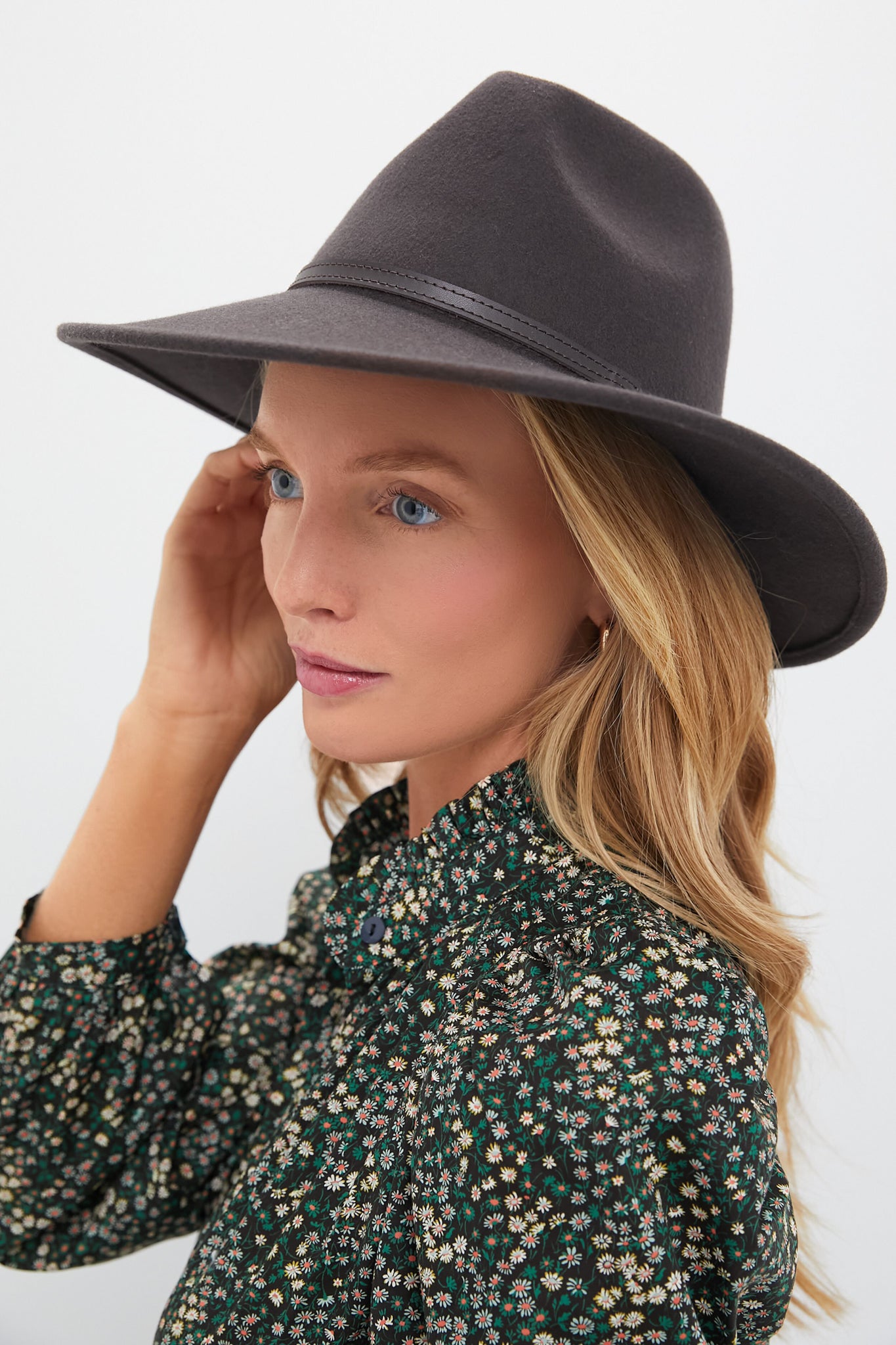 A woman with long blonde hair wears a Barbour fedora and a green, floral-print blouse, looking to the side against a simple background.
