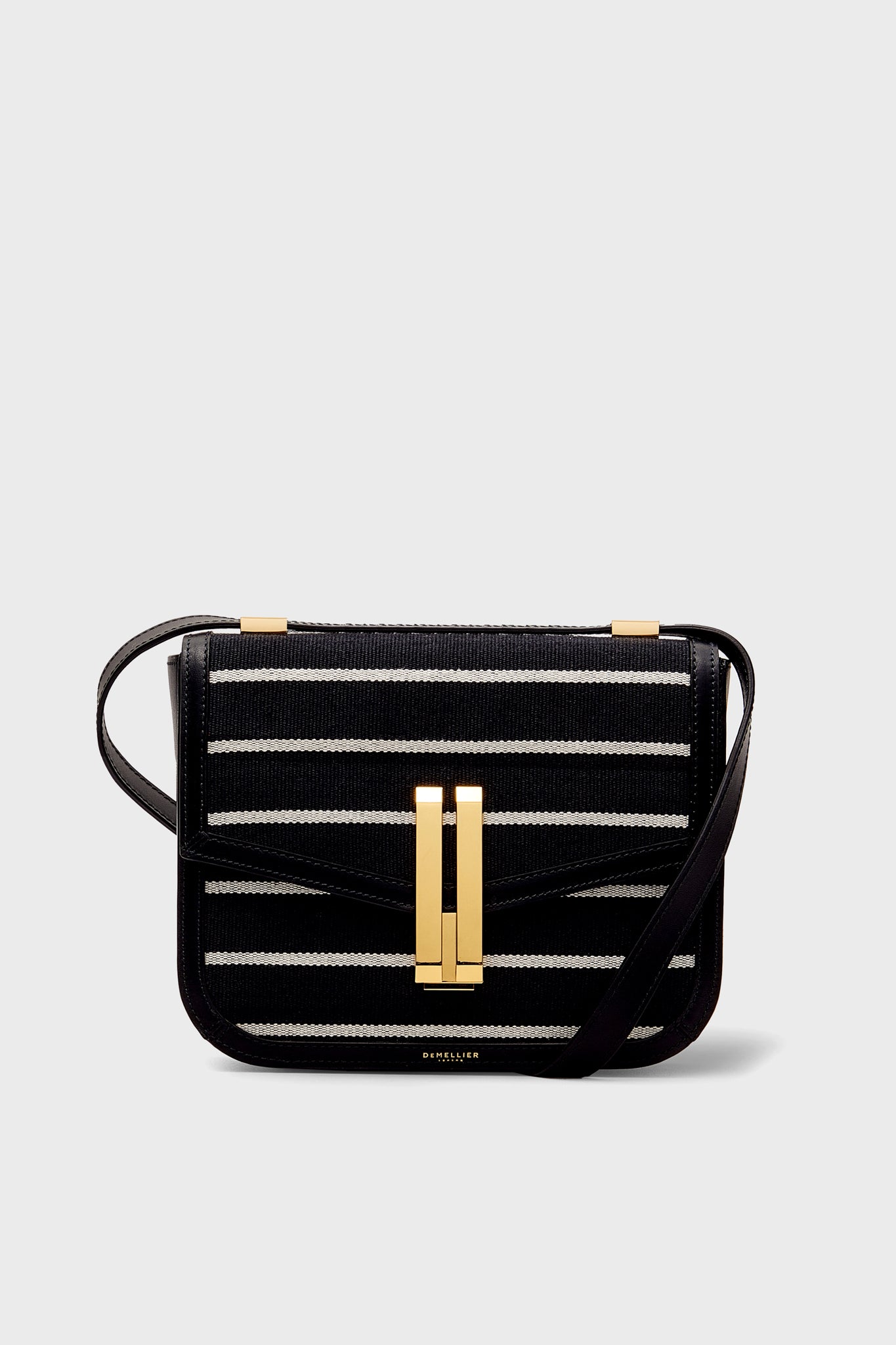 The Demellier Vancouver Bag is a black Breton Stripe Canvas handbag featuring white horizontal stripes, a gold rectangular clasp, and a long black strap—a handcrafted, sustainable accessory for standout style.