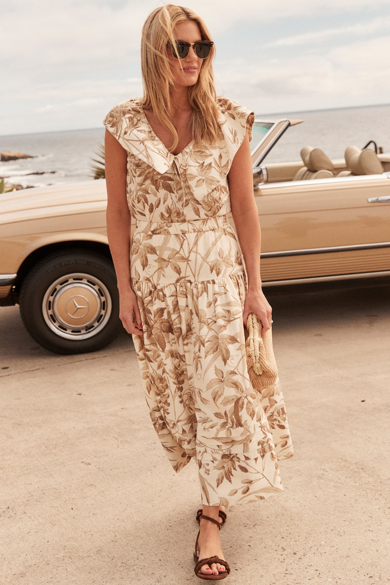 Shea McGee in a brown and white floral printed dress, styled with brown suede sandals and a tan raffia clutch. 