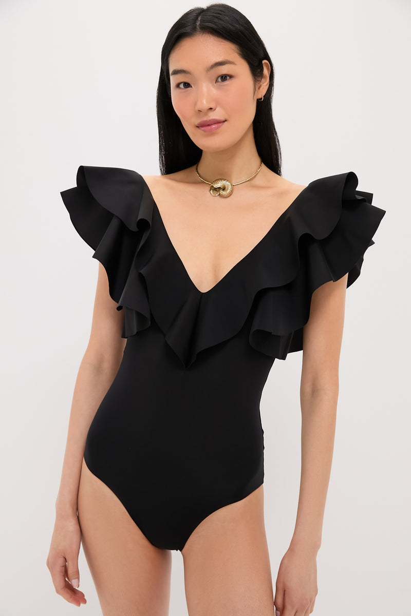 A woman wears the Maygel Coronel Black Santa One Piece, a luxury ruffled swimsuit with a deep V-neck and layered ruffle sleeves, paired with a gold choker necklace.