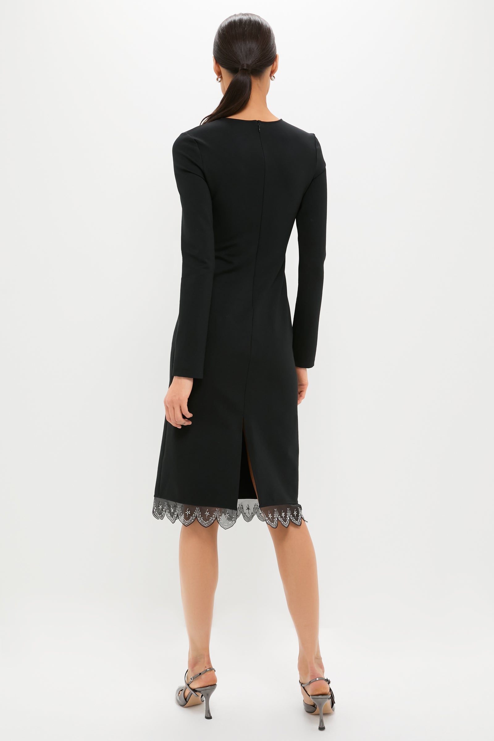 Woman in a classic Black Hunter Dress with scalloped lace hem, effortlessly styled with metallic-grey sandals, embodying high-end, fun elegance against a simple backdrop.